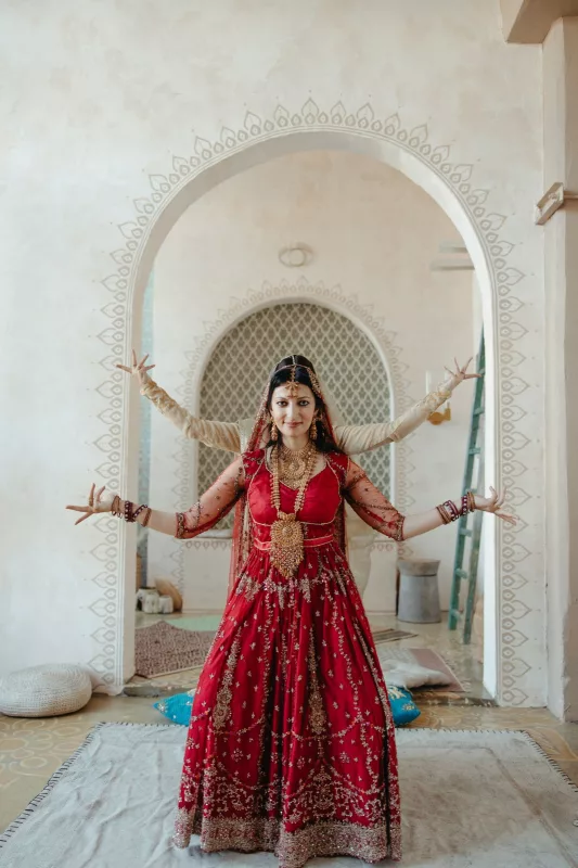 Bride in a red bridal lehenga with gold zari work, standing with arms stretched out under an arched doorway