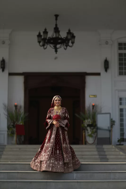 Bride in a maroon velvet bridal lehenga with gold embroidery, standing on palace steps