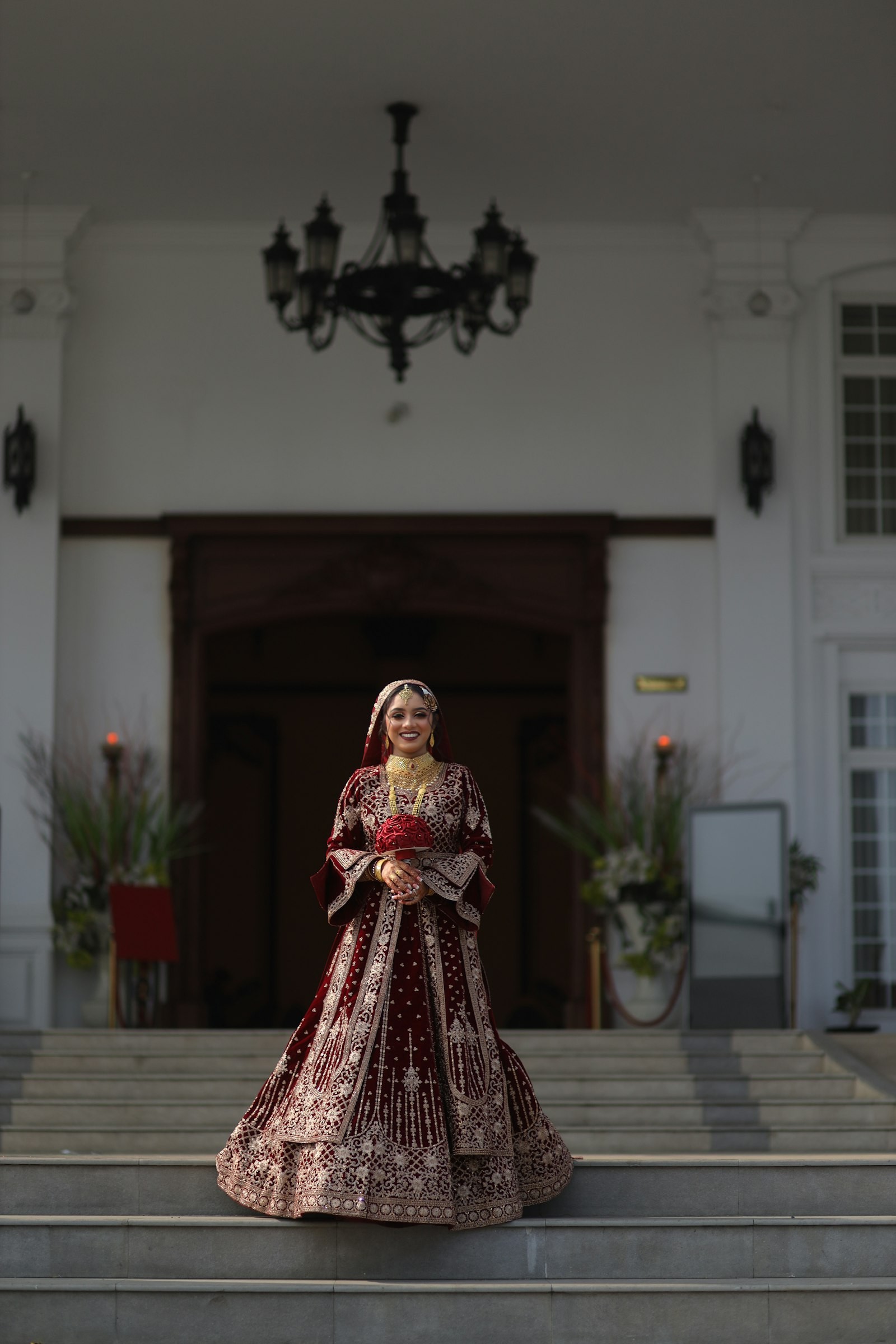 Bride in a maroon velvet bridal lehenga with gold embroidery, standing on palace steps
