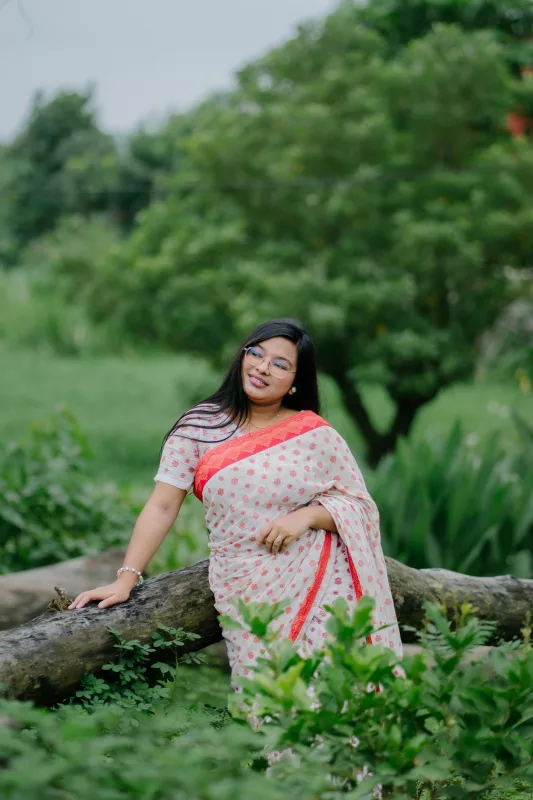 Woman in a white saree with a red border and floral print, standing in a garden