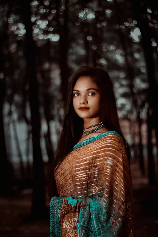 Woman in a copper brown silk blend saree with a teal border, standing among trees