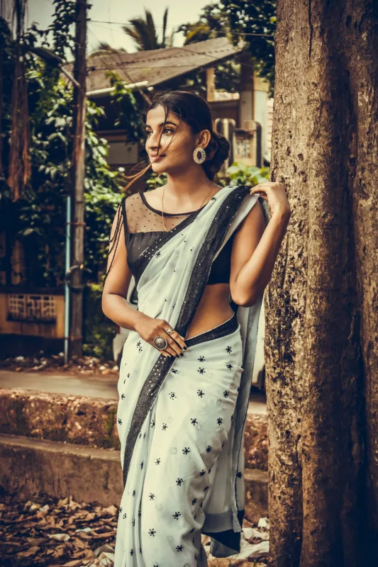 Woman in a pale grey printed saree with a dark border and a black blouse, standing among trees