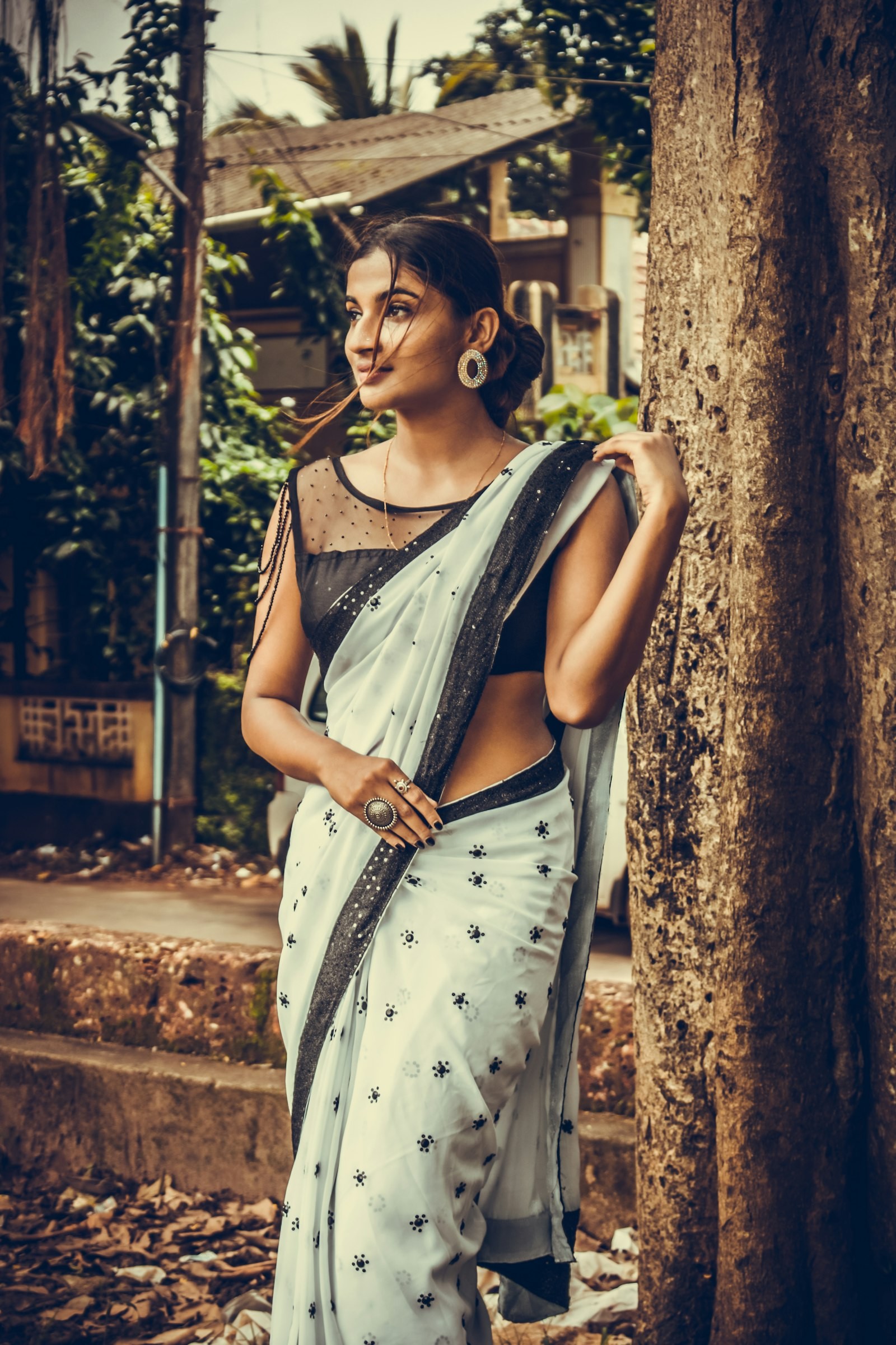 Woman in a pale grey printed saree with a dark border and a black blouse, standing among trees