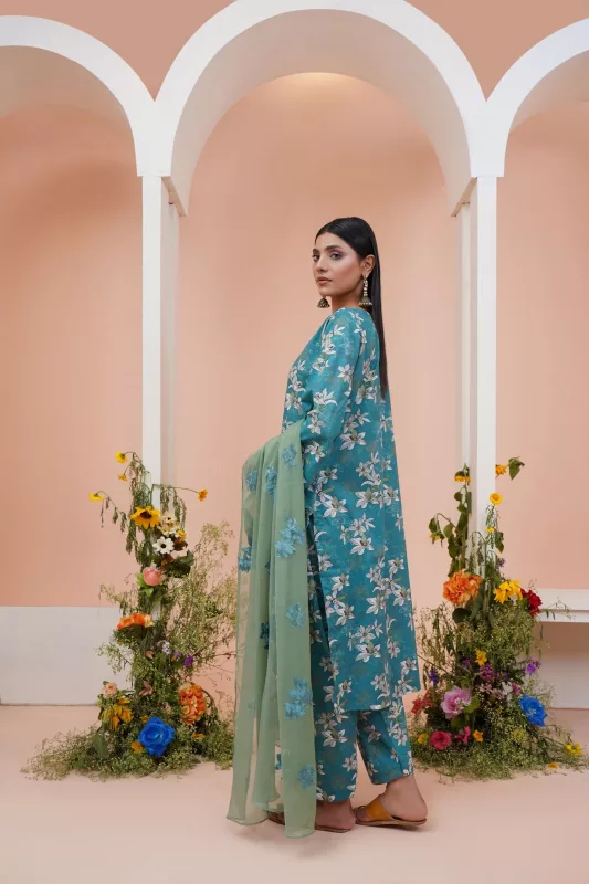 Woman in a teal floral printed straight suit with a sage green dupatta, standing between flowers under pink arches