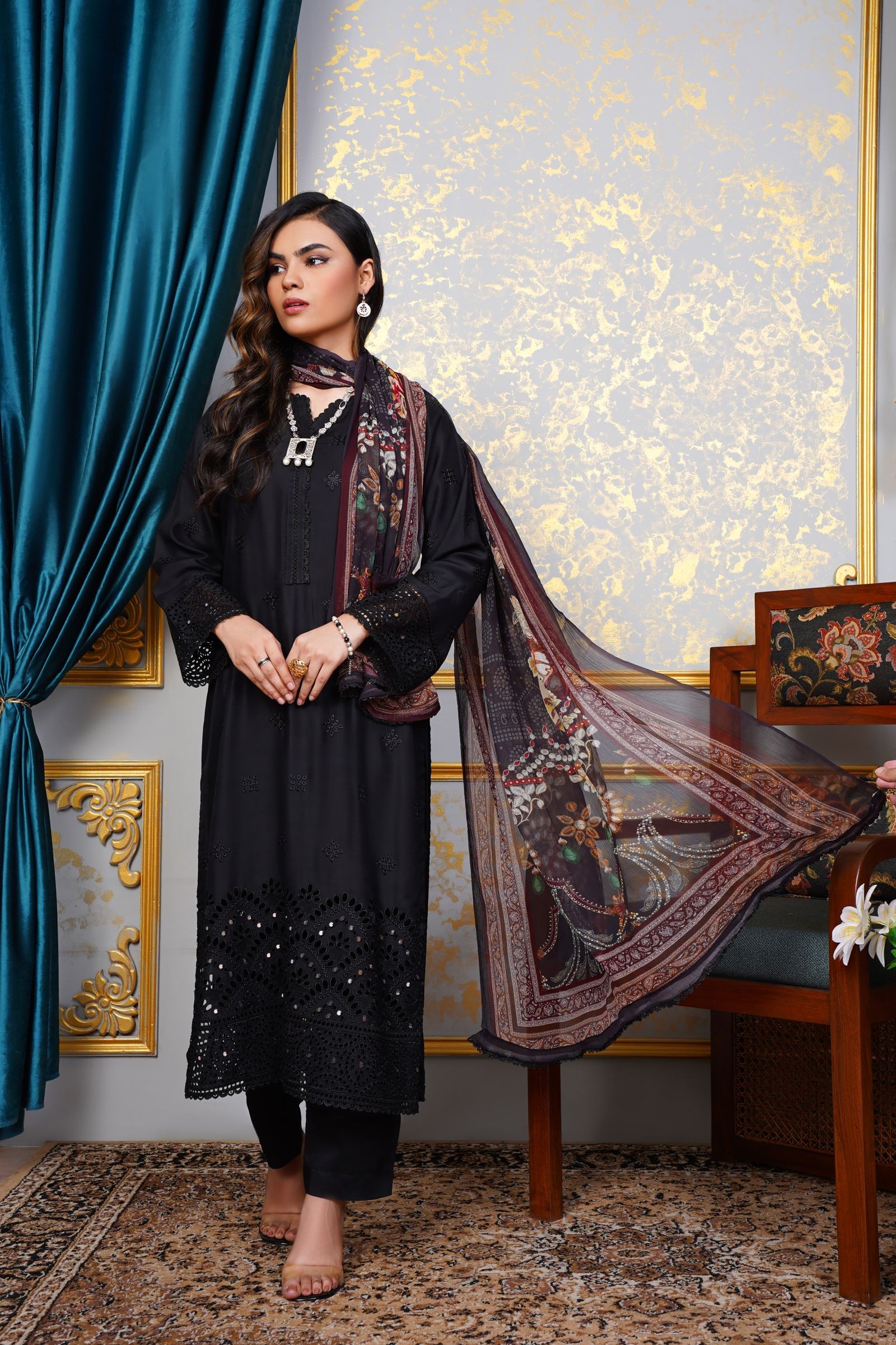 Woman in a black cutwork embroidered straight suit with a printed dupatta, standing in front of a gold patterned wall