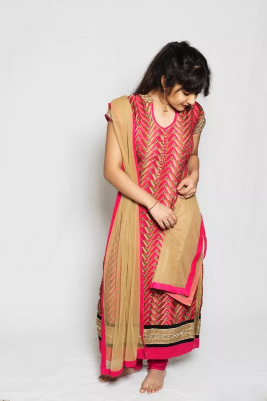Girl in a pink and gold churidar-style suit with a gold net dupatta, standing in a white studio