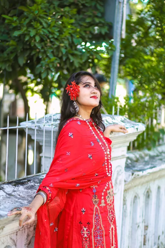 Woman in a red embroidered kurta suit with a matching red dupatta, leaning on a garden wall